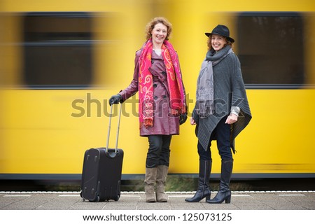 Two female friends are standing on the train station platform with luggage as a yellow train speeds past them in the background