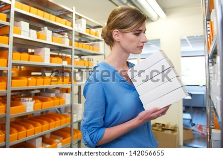 Young female pharmacist holding boxes and looking for medicine on shelves
