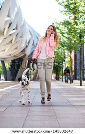 Portrait of a happy young woman walking her dog and talking on cellphone in the city