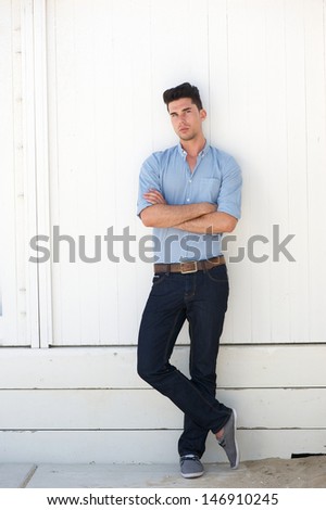 Portrait of an attractive young male fashion model in blue jeans and shirt standing against white wall outdoors
