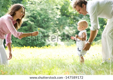 Portrait Of A Happy Young Family Teaching Baby To Walk In The Park