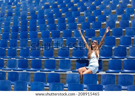 Young woman sitting in stadium cheering with arms raised