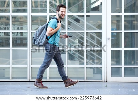 Side view portrait of a young man walking on sidewalk with mobile phone and bag
