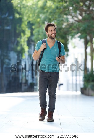 Full body portrait of a cheerful young man walking with mobile phone and bag