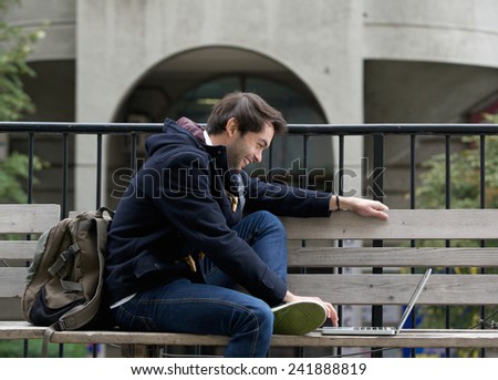 Side view portrait of a young man sitting on bench smiling at laptop
