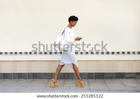 Full length side portrait of a young woman walking and sending text message on cell phone