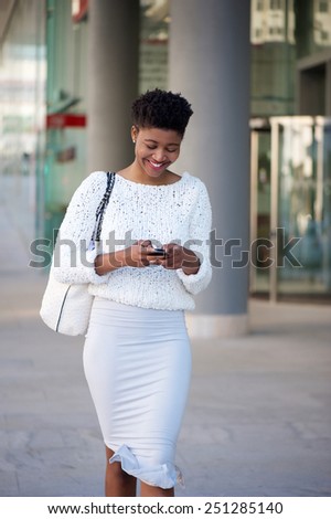Portrait of a cheerful young woman walking on sidewalk and sending text message on mobile phone