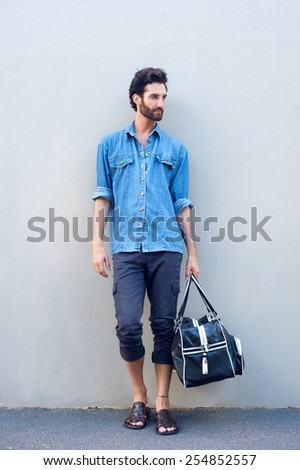 Full body portrait of a male fashion model with beard holding a travel bag on gray background