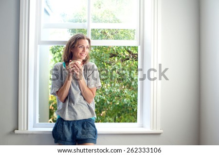 Portrait of a happy woman standing by window smiling with cup of coffee