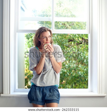 Portrait of a healthy woman standing by window at home with a cup of coffee