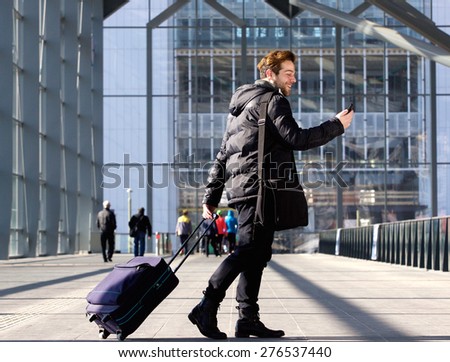 Portrait of a young man walking with suitcase and looking at mobile phone