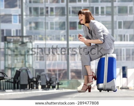 Side portrait of a business woman sitting on suitcase and laughing with mobile phone