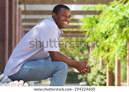 Close up portrait of a smiling relaxed young black guy sitting on bench in the park