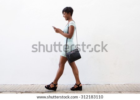 Full body side portrait of a smiling african american young woman walking and looking at cell phone