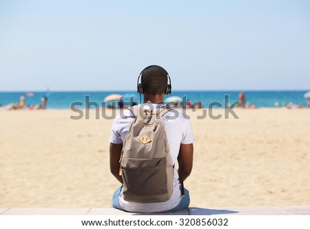 Portrait from behind of a young man sitting by the beach listening to music on headphones