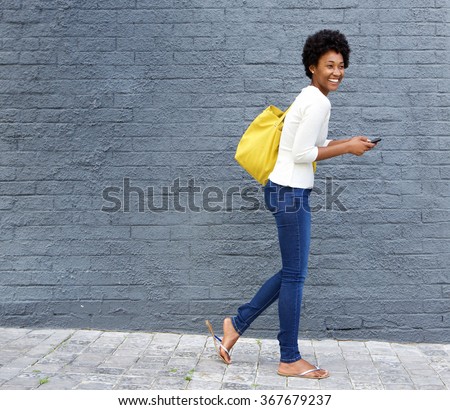 Full length portrait of a cheerful young african woman walking on street with a mobile phone