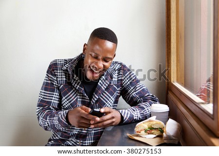 Portrait of smiling young african man sitting at a cafe and reading text message on his cell phone