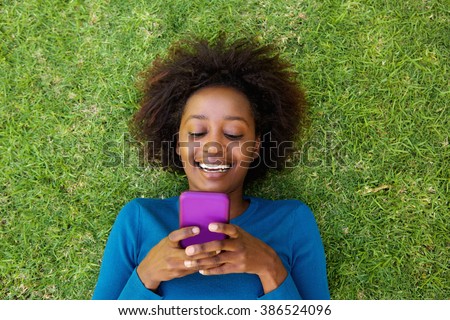 Portrait from above of a smiling african woman lying on grass looking at cell phone