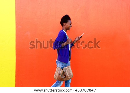 Side view portrait of trendy young black woman walking and  reading text message on her mobile phone