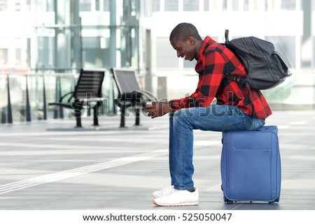 Side portrait of happy young man sitting on traveling bag and looking at cell phone