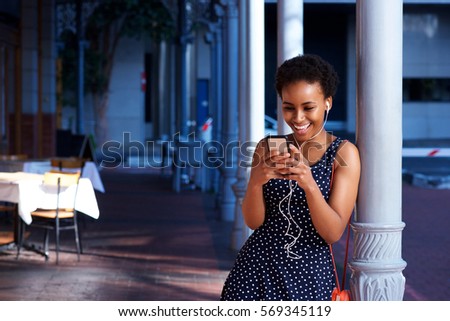 Portrait of smiling young black woman with earphones looking at cellphone