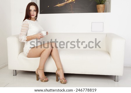 Girl in white dress with a cup of coffee in hand sitting on a white sofa