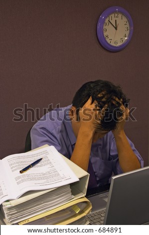 Stress At Work - A man with his head down clenching his hair while working late into the night. He is surrounded by his computer & piles of documents, and the clock shows close to midnight.