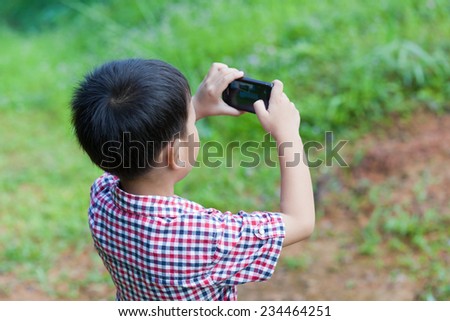 Portrait of little asian boy taking photos by digital camera on smartphone with good intentions, outdoor in the midst of glade on holiday