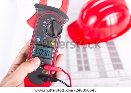 Hand of technician holding digital clamp multimeter over blueprint and red hard hat background