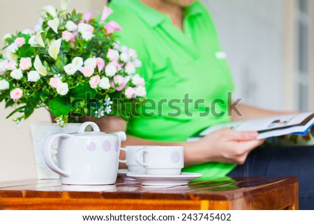 Young woman reading magazine. white porcelain set for tea or coffee and vase of artificial flowers on wooden table at home exterior in relax time, shallow DOF teapot in focus