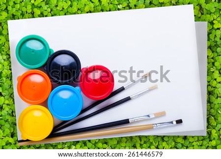 Multicolored drawing instruments (watercolor paints, paintbrush, blank sheet of paper) for create imagination over green clover background, creativity concept. Top view