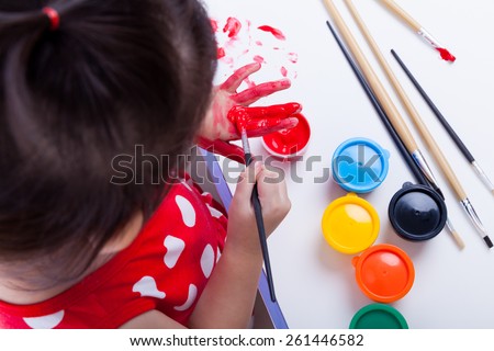 Little asian (thai) girl painting her palms using multicolored drawing instruments (watercolor paints, paintbrush), creativity concept, studio shot, top view