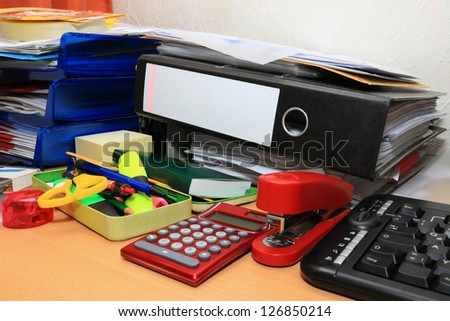 A messy desktop with stacks of files and other documents, all kind of office supplies and part of a keyboard.