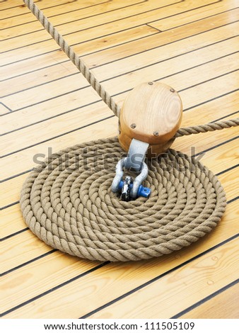 A photo of a thick rope in spiral or ring shape on a wooden sailing ship floor and another rope going through pulley which is attached with shekel into the floor