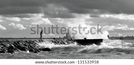 Human standing on a pier or quay in a stormy and turbulent sea at a gloomy but but sunny day. City silhouette of Tallinn in background.