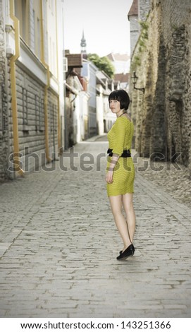 Young woman in short yellowish or greenish knitted dress walks and look back on a street in the Old Town of Tallinn, Estonia.