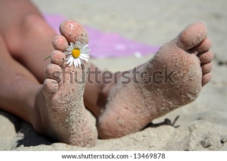 Holiday concept of two feet on a beach chair against white sandy beach and sunny blue sky.