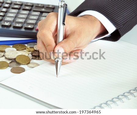 Businessman hand holding a pen writing on empty paper with coins & calculator aside - business & financial concept