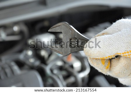 Hand wearing glove holding wrench in front of car engine background