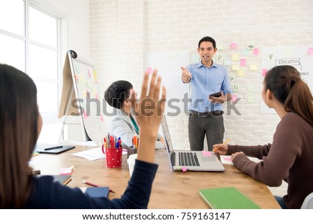 Asian teacher pointing hand to one of female college student raising her hand to ask a question in classroom