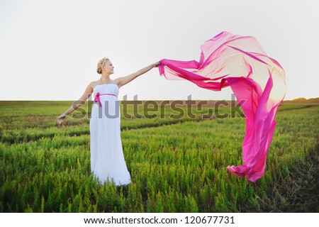 young woman in a white greek dress stands in field holds pink cloth