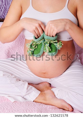 young pregnant woman sitting on bed at home, holding a tiny socks