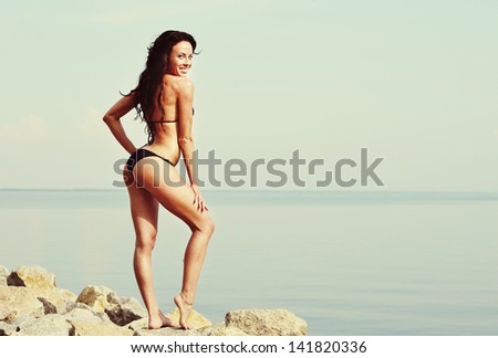 Caucasian young brunette wearing bikini stands on stone next to sea