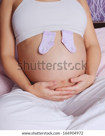 young pregnant woman sitting on bed at home, holding tiny socks for a baby