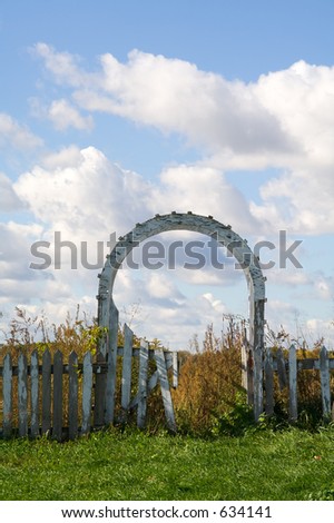 A dilapidated picket fence and gate lead to blue skies and grassy pastures beyond.