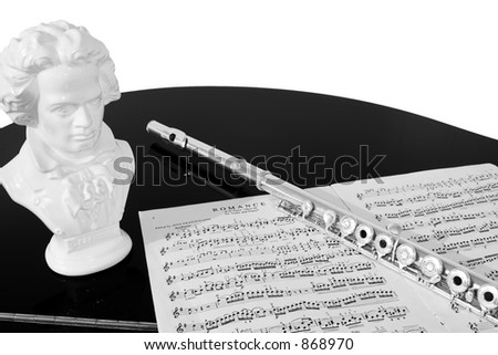 A flute, musical score and bust of Beethoven rest on a black piano top, isolated against a white background. Black and white (grayscale).