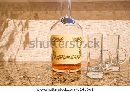 Beautiful cognac bottle with amber liquid, and two fancy shot glasses, on a granite counter set against travertine backsplash.