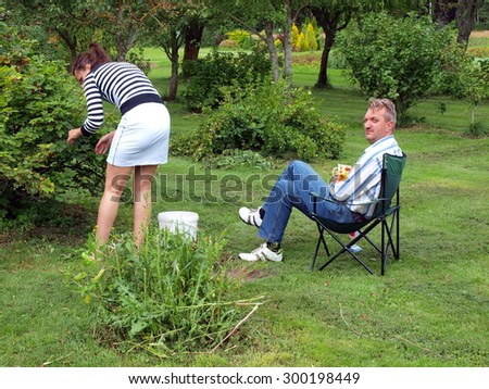 In garden young woman is harvesting currants and man sits in chair drinking coffee look to her back.