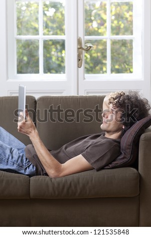 A man reclines on a sofa reading from a tablet computer.