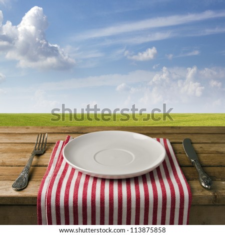 Empty Plate With Fork And Knife On Wooden Table Against Blue Sky And Meadow. Table Arrangement.